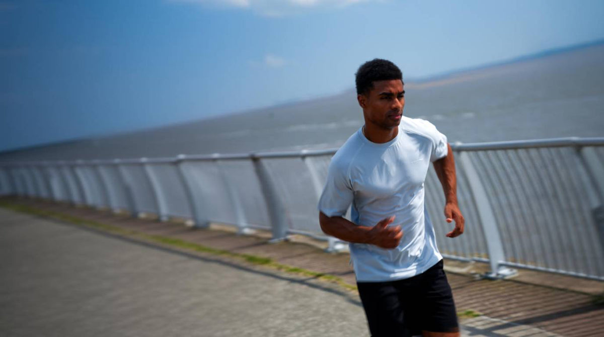 A man jogging outdoors on a paved path next to a railing with water and sky in the background, wearing a white athletic t-shirt and black shorts.