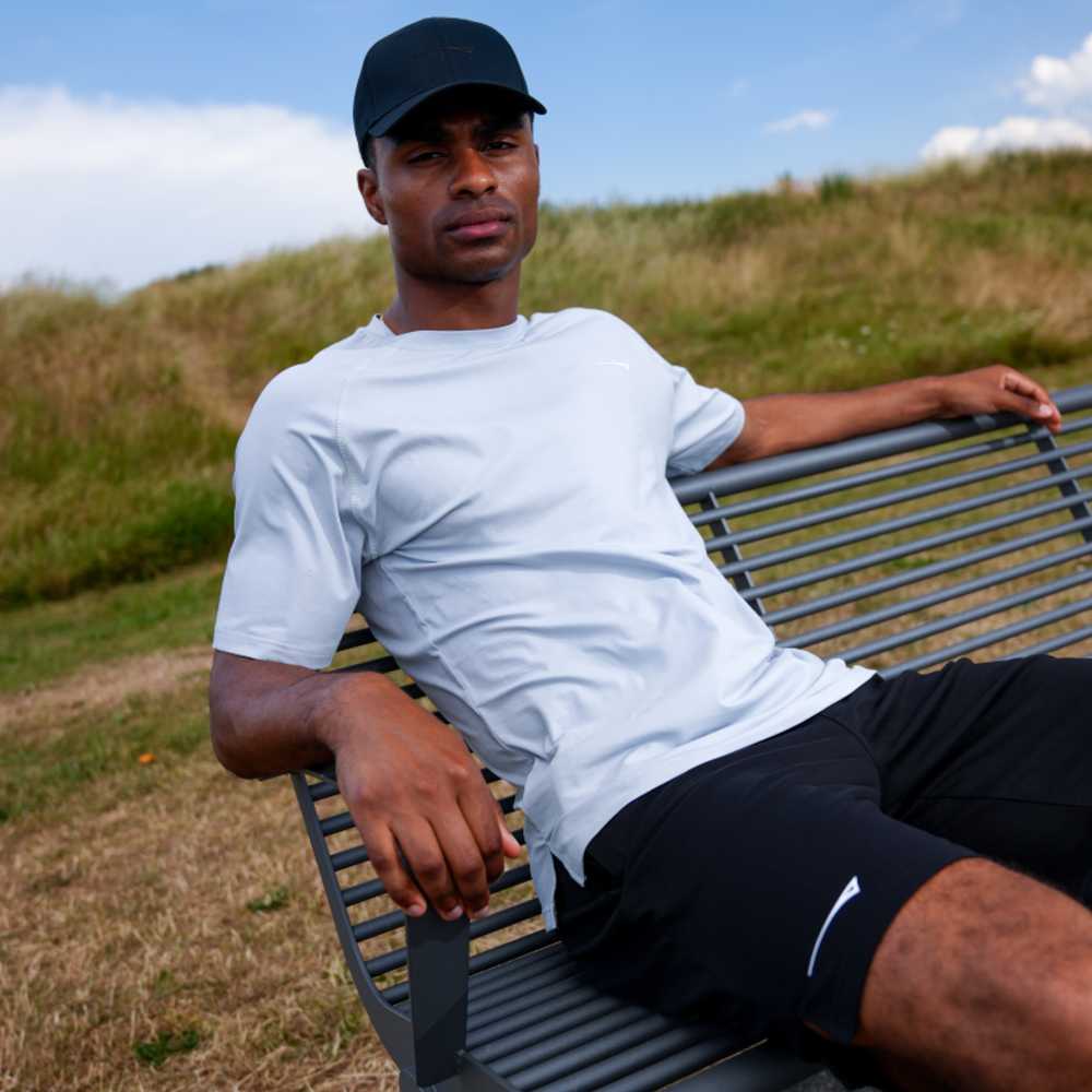 A man wearing a light gray athletic t-shirt, black shorts, and a black cap sits on a metal park bench outdoors with grassy hills in the background.
