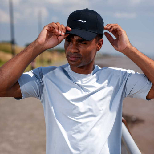 A man wearing a light blue athletic t-shirt and a black baseball cap with a white logo, adjusting his cap outdoors on a sunny day.