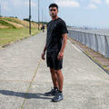 A man wearing a black athletic t-shirt and matching black shorts stands on a paved path next to the sea under a blue sky.