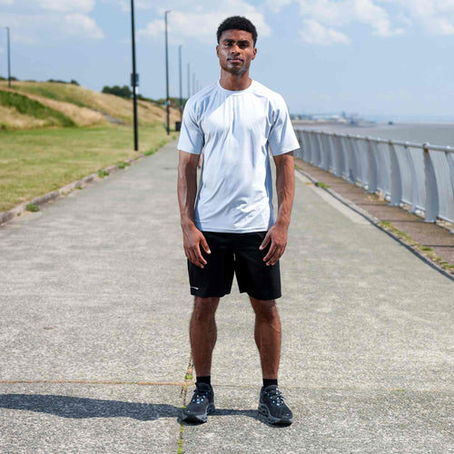 A man stands on a paved path outdoors near the water, wearing a white athletic t-shirt, black athletic shorts, and dark running shoes.